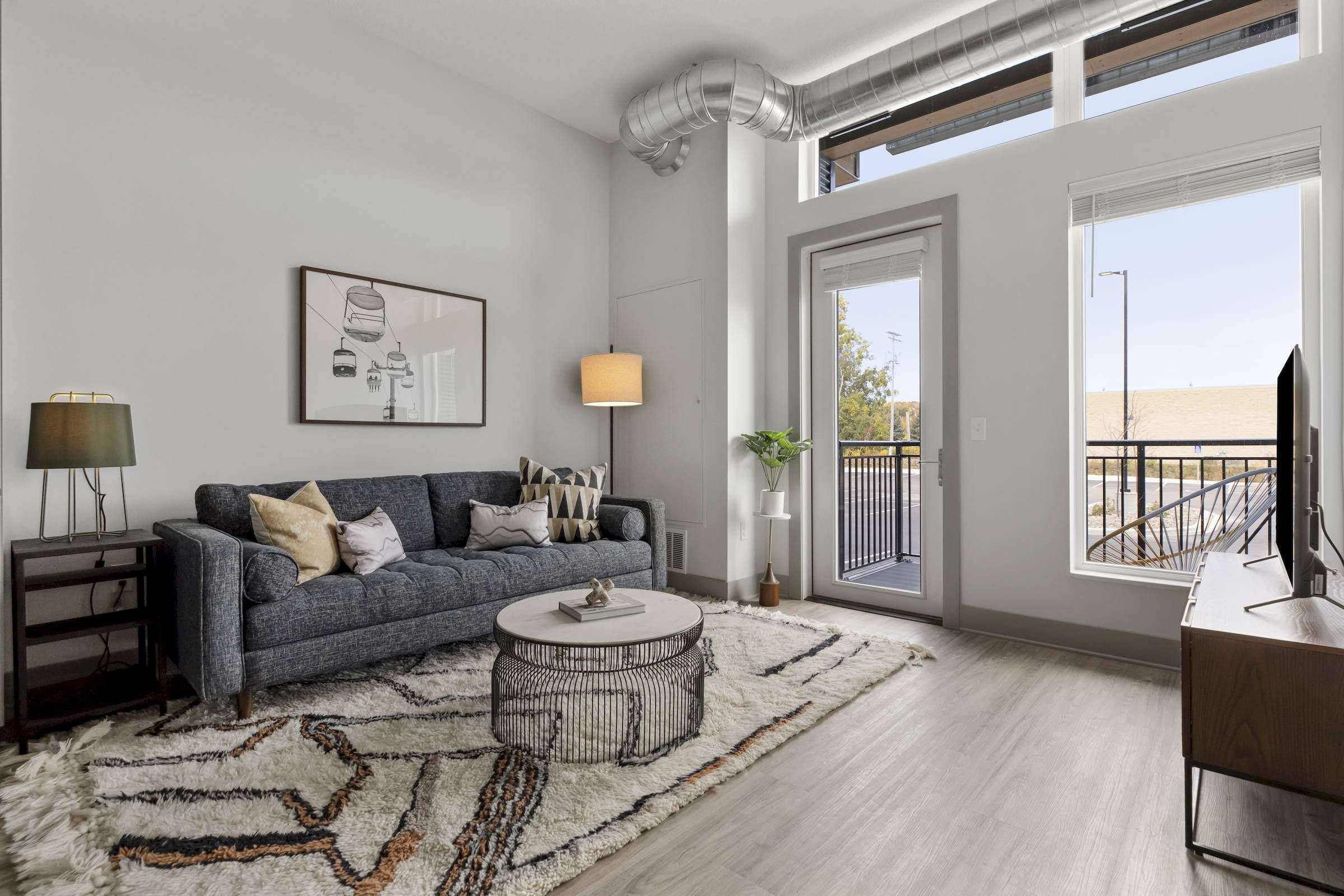 Modern living room with a gray sofa, round coffee table, rug, and large windows letting in natural light.