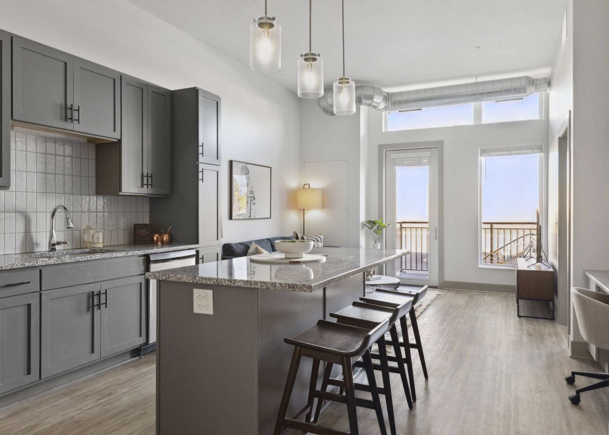 Modern kitchen with gray cabinets, island seating, pendant lights, and large windows letting in natural light.