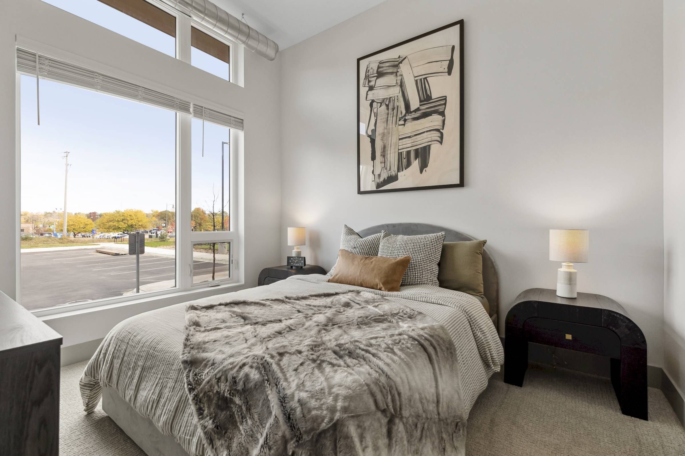 Modern bedroom with large window, neutral bedding, abstract wall art, and two nightstands with lamps.