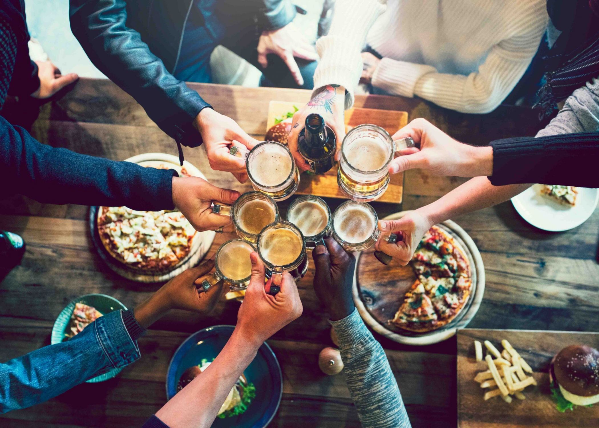 People clinking beer mugs over a wooden table with pizza and fries, viewed from above.