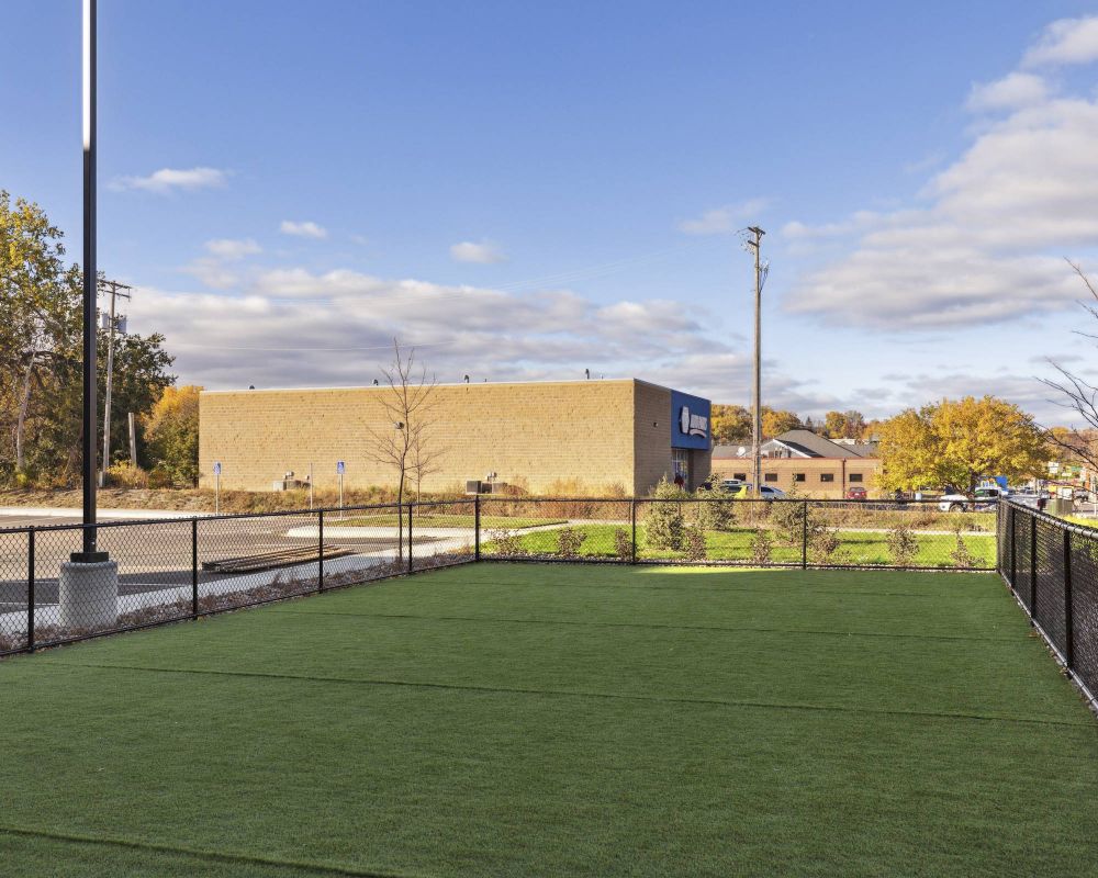 Fenced grassy area next to a parking lot, with buildings and trees in the background under a partly cloudy sky.